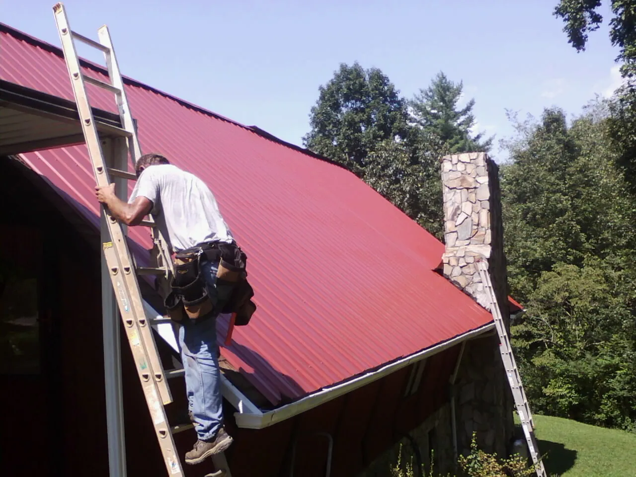 Roofer on a ladder beside a red metal roof and stone chimney — Soffit & Fascia Repair by Roofing Brothers in Susan
